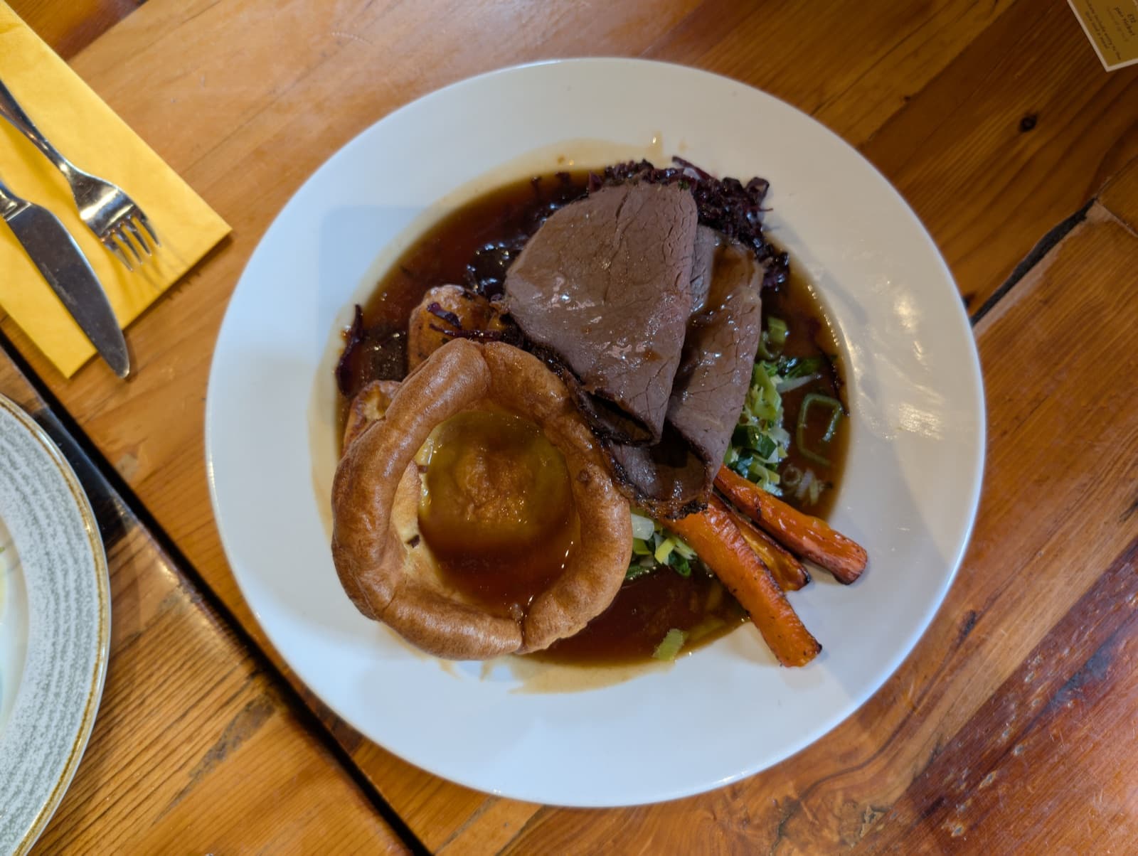 A plated roast beef dinner on a wooden table: sliced beef in gravy with a Yorkshire pudding, a roast potato, carrot and red cabbage.